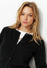 Cardigan noir en matériau doux, avec un col rond, une fermeture à boutons sur le devant et des manches longues, superposé sur une chemise blanche.