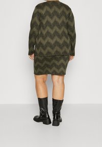 Černé kotně z bot s vysokým podpatkem a masivním podpatkem, kombinované se zelenými šaty s vlnitým vzorem, pohled zezadu.