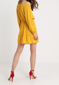 Ärmelloses gelbes Kleid mit langen Ärmeln, gerafftem Bund und Volant-Saum. Kombiniert mit roten High-Heels.