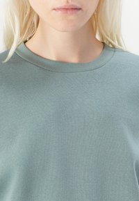 Šviesiai žalia pūkinė palaidinė su apvaliu kaklu ir švelnia tekstūra, matomi siuvimo detalės ir be aiškių raštų. Artimas vaizdas nuo pečių iki viršaus.