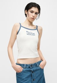 Νεαρό άτομο με κοντά, σκουρόχρωμα μαλλιά, που φοράει λευκό crop top με μπλε λουριά και το κείμενο "Dickies", συνδυασμένο με μια μπλε τζιν φούστα με κουμπιά.