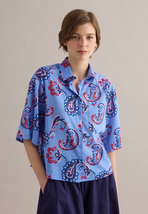 Jeune femme portant une chemise bleue ample à boutons avec un motif cachemire rouge et bleu marine, associée à un pantalon large foncé, debout avec la main dans la poche.
