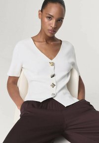 Top bianco a maniche corte con scollatura a V profonda e bottoni decorativi in oro, argento e cristalli. Indossato con pantaloni marrone scuro.