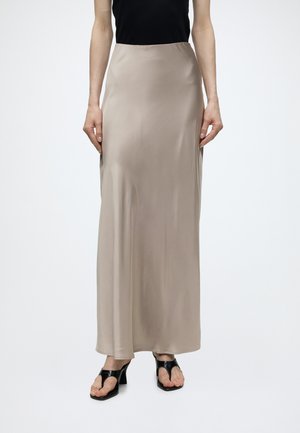 Jupe longue en satin beige portée avec un haut noir sans manches et des sandales à talons ouvertes noires sur un fond blanc uni.