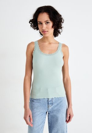 Top de tirantes acanalado verde claro con encaje en el escote y los tirantes. El dobladillo es con forma de ondas. Combinado con unos vaqueros de tiro alto azul claro.