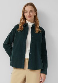Chemise en velours côtelé vert foncé avec un devant à boutons, des manches longues et une coupe décontractée, associée à un col roulé blanc et un pantalon beige.