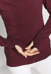 Maroon zip-up hoodie lavet af blødt stof, med tommelfingerhuller i ærmerne, ribstrikkede manchetter og en glat lynlås.