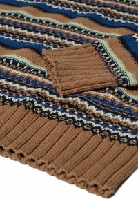 Brūns adīts džemperis ar daudzkrāsainām zigzag svītrām. Tam ir ribotas piedurknes un apakšmala, kā arī sarežģīti ģeometriski raksti zilā un krēmkrāsā.