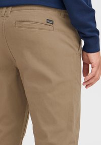 Khaki chinos i en mjuk bomullsblandning, med en bakficka och en liten svart logotagg. Tyget är slätt med en matt finish.