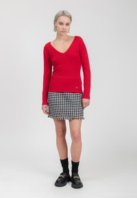 Roter figurbetonter Pullover mit V-Ausschnitt, kombiniert mit einem schwarz-weißen Hahnentrittmuster-Rock mit Fransenverzierung. Schwarze Platform-Loafer mit Ketten.