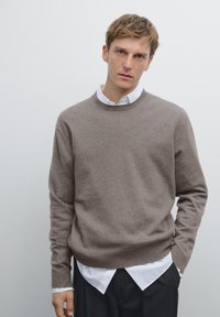 Brauner Strickpullover mit rundem Halsausschnitt, über einem weißen Hemd mit Kragen getragen. Weiche Textur, lockere Passform und lange Ärmel.