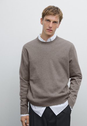 Pull en tricot marron à col rond, porté par-dessus une chemise blanche à col. Texture douce, coupe décontractée et manches longues.
