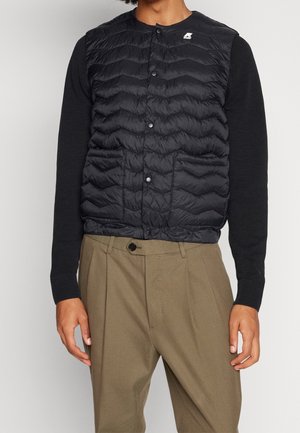 Mann trägt schwarze wattierte Weste über schwarzem Langarmpullover und hellbraunen, plissierten Hosen, steht vor einfarbigem Hintergrund.