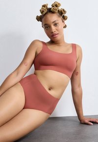 Kvinna med rullat blont hår sitter lutad bakåt på en arm, iförd matchande rostfärgad sömlös bh och högmidjade trosor.