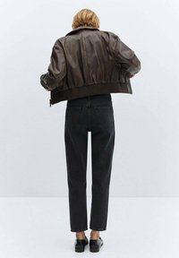 Persona vista de espaldas vistiendo una chaqueta de cuero marrón, jeans negros de tiro alto y mocasines negros, contra un fondo claro y simple.