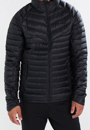 Schwarze gesteppte Steppjacke mit Frontreißverschluss, getragen von einer Person mit entspannt hängen gelassenen Händen an den Seiten, vor neutralem Hintergrund.