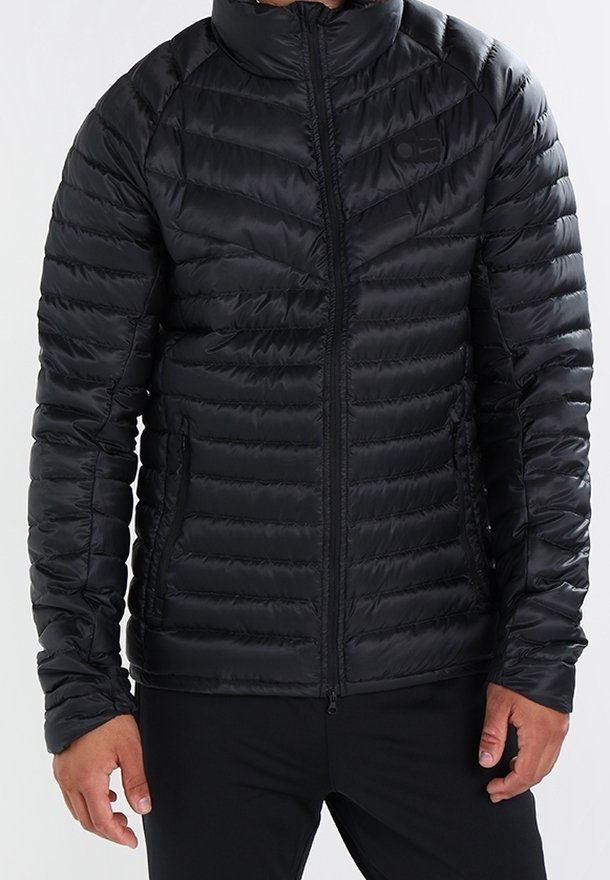 Veste matelassée noire à capuche avec fermeture éclair à l'avant, portée par une personne les mains détendues sur les côtés, sur un fond neutre.