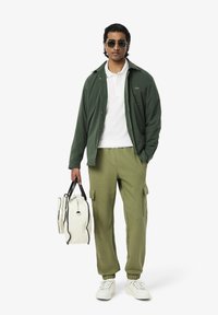 Chemise verte à boutons sur un polo blanc, accompagnée de joggers olive et de baskets blanches. Un sac blanc avec des accents noirs est tenu à la main.