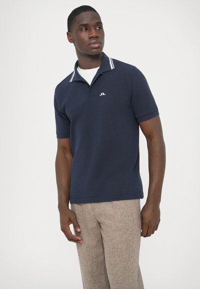 Tmavomodré polokošeľa s bielym pruhovaným golierom, krátkymi rukávmi a vyšívaným logom na hrudi; textúrovaná látka v kombinácii s béžovými vzorovanými nohavicami.