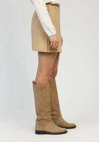 Bottes en daim beige à hauteur de genou avec semelle plate et bout arrondi, assorties à une mini-jupe beige et un haut à manches longues blanc.
