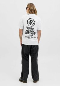 Person med langt hår iført solbriller, en hvid UFC "Ultimate Fighting Championship" T-shirt, sorte bukser og sorte støvler, stående med ryggen til.