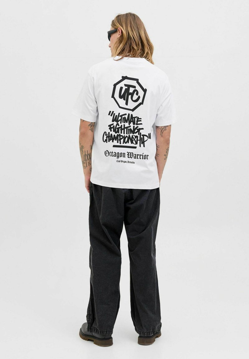 Person med langt hår iført solbriller, en hvid UFC "Ultimate Fighting Championship" T-shirt, sorte bukser og sorte støvler, stående med ryggen til.