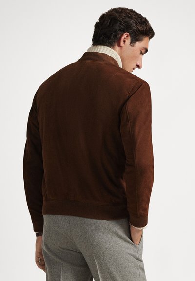 Rjava semiš jakna z rebrastim ovratnikom in manšetami, mehke teksture in sproščenega kroja, v kombinaciji s svetlo sivimi hlačami.