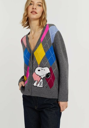 Vrouw die een grijze cardigan draagt met kleurrijke ruitpatronen en een Snoopy cartoonfiguur op de voorkant, gecombineerd met donkere spijkerbroek.