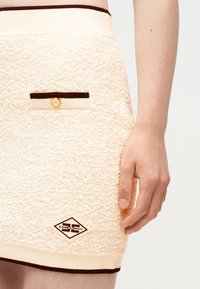 Κοντινό πλάνο χεριού που ακουμπά δίπλα σε φούστα με κρεμ υφή, σκοτεινό περίγραμμα, μικρή τσέπη και ετικέτα εμπορικού σήματος σε σχήμα διαμαντιού κοντά στο τελείωμα.