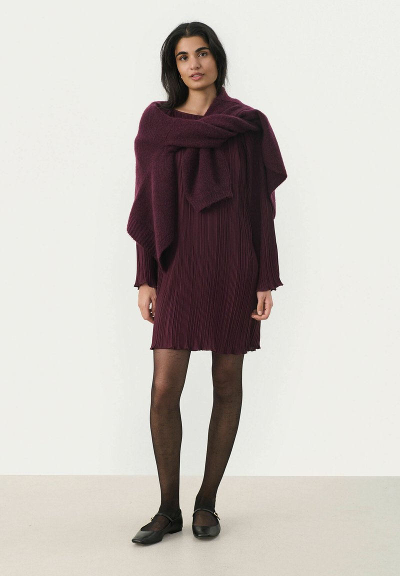 Pflaumenfarbendes, gefältetes Kleid mit langen Ärmeln, kombiniert mit einem passenden gestrickten Schal. Getragen mit schwarzen Strumpfhosen und flachen Schuhen, die ein schlichtes Design aufweisen.