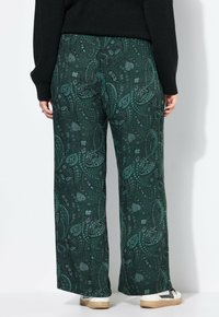 Personne portant un pantalon large à motif paisley vert foncé, un pull noir et des baskets blanches avec des détails noirs, se tenant contre un mur blanc.
