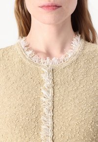 Strikkegenser i lys beige med strukturert stoff og en frynsete hvit krage. Har knappelukking foran.
