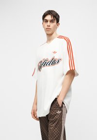 Młody mężczyzna ubrany w białą koszulkę Adidas z pomarańczowymi paskami i logo, w połączeniu z brązowymi spodniami dresowymi Adidas w wzory, stojący z ręką w kieszeni.