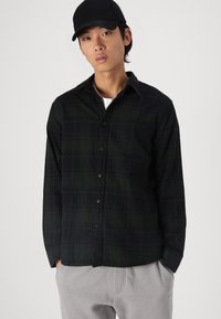 Black and green plaid button-up shirt in a soft fabric, featuring a collar, long sleeves, and black button closures. Paired with gray pants.