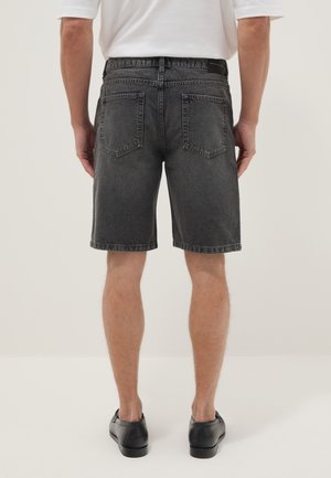 Man draagt zwarte denim shorts, wit T-shirt en zwarte schoenen, staand met zijn rug naar voren tegen een effen lichte achtergrond.