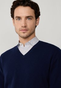 Granatowy sweter z dekoltem w serek, wykonany z miękkiej dzianiny, noszony na białej koszuli w cienkie niebieskie paski, z klasycznym stylem kołnierzyka.