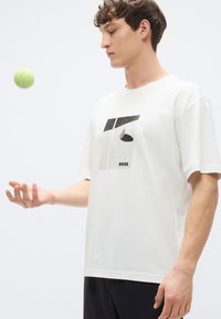 Λευκό βαμβακερό t-shirt με γραφικό σχέδιο γεωμετρικών σχημάτων και λογότυπο «BOSS». Το μοντέλο κάνει ζογκλερικά με μια μπάλα τένις.