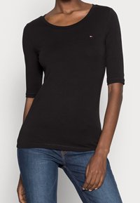 Černé bavlněné tričko s třičtvrtinovými rukávy, kulatým výstřihem a malým logem na hrudi. V kombinaci s modrými džínami.