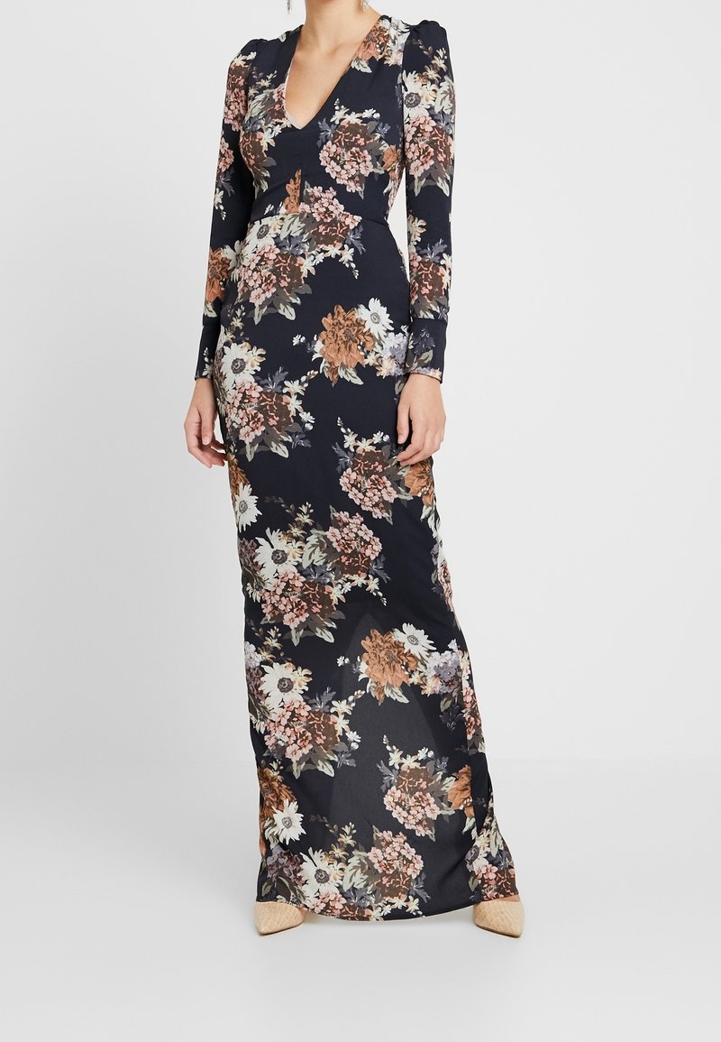 Woman wearing a long dark floral dress with a V-neck and long sleeves, paired with beige quilted pointed shoes.
