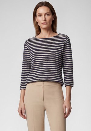 Woman wearing a navy and white striped three-quarter sleeve top and beige high-waisted pants, standing against a plain background.