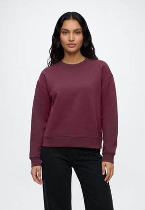 Kvinde med langt mørkt hår iført en ensfarvet bordeaux crewneck-sweatshirt og sorte bukser, står mod en hvid baggrund.