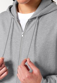 Felpa grigia con zip e cappuccio regolabile, caratterizzata da hardware argentato e una texture morbida. Indossata sopra una t-shirt bianca, con le mani che tengono i lacci.