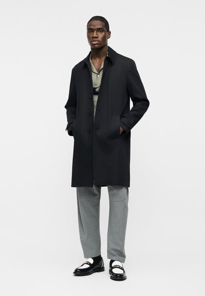 Άνδρας με μαύρο παλτό, πουκάμισο με σχέδια, γκρι παντελόνι και ασπρόμαυρα μοκασίνια, στέκεται μπροστά σε απλό ανοιχτό φόντο.