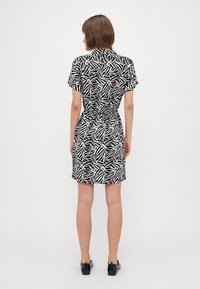 Kratka haljina s kratkim rukavima koja se ističe apstraktnim uzorkom u crno-bijeloj boji, s pojasom na struku i duljinom iznad koljena, izrađena od laganog materijala.