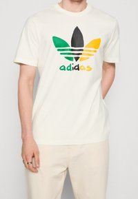 Krémové bavlněné tričko s potiskem černého, zeleného, žlutého a oranžového loga adidas, krátkými rukávy a kulatým výstřihem.
