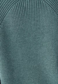 Strickpullover in gedämpftem Blaugrün mit geripptem Textur und gebogenen Schulterdesign. Der Stoff wirkt weich und zeigt feine Nähdetails.