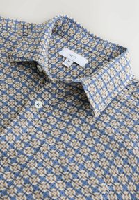 Chemise à carreaux bleue et beige tissée, col classique, boutonnée sur le devant, avec une étiquette indiquant "NEXT" et la taille "M". Texture douce.