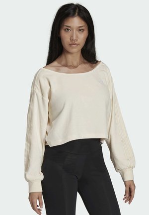 Gekürzter cremefarbener Hoodie mit weitem Ausschnitt, langen puffigen Ärmeln, gerippten Bündchen und dezentem Logo-Detail auf der Brust. Kombiniert mit schwarzen Leggings.