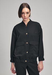 Zwarte jas met een hoge kraag, twee voorzakken en gouden knopen. Heeft een gerimpelde zoom en een gladde textuur. Geschikt voor casual kleding.