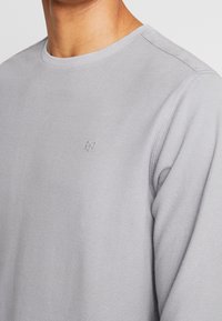 Graues Langarmshirt mit rundem Ausschnitt, aus strukturiertem Stoff und einem kleinen gestickten Logo auf der Brust. Glattes, figurbetontes Design.